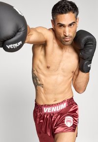 Melni boksa cimdi ar "Venum" logotipu, sarkanas satīna šorti ar baltiem akcentiem un logotipu. Modelim ir tetovējums uz kreisās rokas.