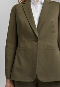 Veste vert olive avec fermeture à un bouton, revers crantés et deux poches horizontales sur une chemise blanche boutonnée.