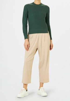 Kvinde iført en mørkegrøn ribstrikket sweater, beige højttaljede bukser med vidde i benene og hvide sneakers, stående foran en ensfarvet baggrund.