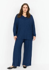 Sötétkék otthoni viselet szett, laza V-nyakú felsővel és bő szárú nadrággal, puha textúrájú anyagból készült. Hozzávaló fekete lapos cipők.