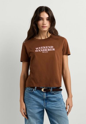 Donna con lunghi capelli castani che indossa una maglietta marrone con la scritta "WEEKEND WANDERER" e jeans blu con cintura nera, in piedi davanti a uno sfondo semplice.