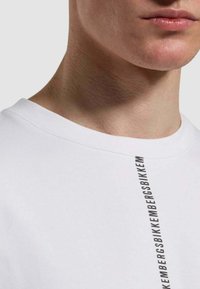 T-shirt bianco in cotone con scollo rotondo e un design testuale verticale nero sul lato. Tessuto morbido e vestibilità standard.