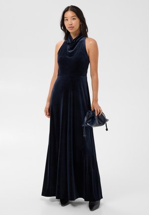 Femme portant une robe maxi sans manches en velours bleu foncé, tenant une pochette assortie à cordon, debout devant un fond uni.