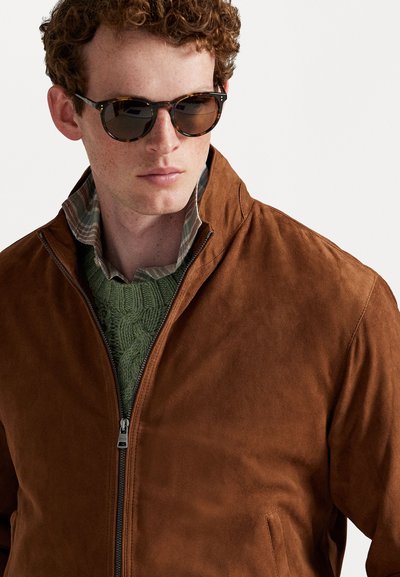 Braune Wildlederjacke mit Stehkragen und durchgehendem Reißverschluss. Über einem grünen Strickpullover und einem karierten Hemd getragen, ergänzt mit Sonnenbrille.