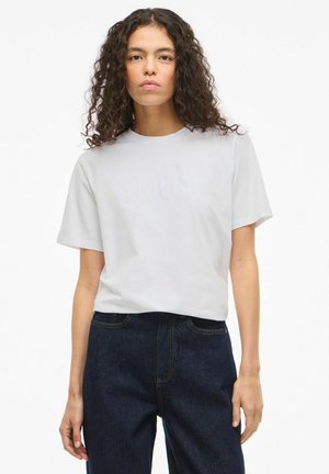 Lichtgrijs katoenen T-shirt met korte mouwen, met in het midden "CHÉRI" in reliëf, gecombineerd met donkerblauwe spijkerbroek.