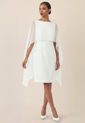 Frau trägt ein weißes knielanges Kleid mit transparenten Umhangärmeln, kombiniert mit weißen spitzen High Heels, steht vor einem schlichten beigen Hintergrund.