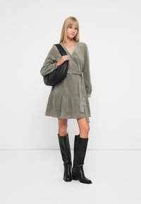 Kort, olivengrønn kordfløyelskjole med lange ermer, belted midje og volangkant. Brukt med knelangne svarte lærstøvler og en svart veske.