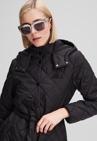 Veste matelassée noire avec capuche, dotée de boutons-pression et d'une taille ceinturée. Tissu texturé avec un logo brodé sur la poitrine.