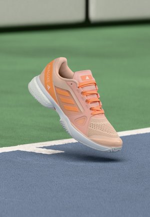 Lichtroze en oranje Adidas Avaluxe tennisschoen zweeft boven een groen-blauw veld vlakbij een witte lijn.