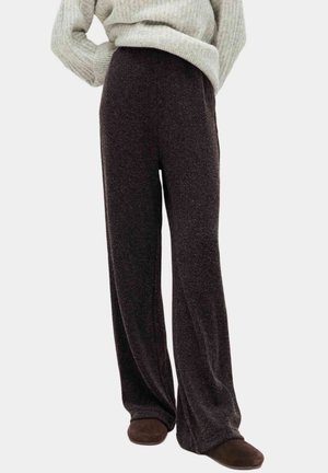 Person trägt dunkelbraune Hosen mit weitem Bein, hellgrauen Strickpullover und dunkelbraune Slipper vor einfachem Hintergrund.