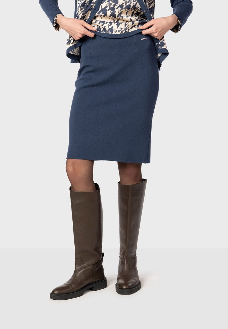 Jupe bleu marine à genoux en tissu extensible, associée à des bottes en cuir marron, hauteur genou, avec un petit talon et un design sobre.