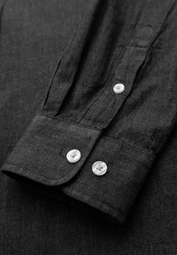 Gros plan d'un poignet de chemise noire à manches longues avec deux boutons blancs et une partie de la patte de manche visible sur le tissu.