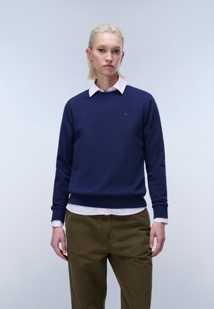 Jeune personne aux cheveux blonds courts portant un sweat-shirt bleu marine par-dessus une chemise blanche à col et un pantalon vert olive, debout devant un fond uni.