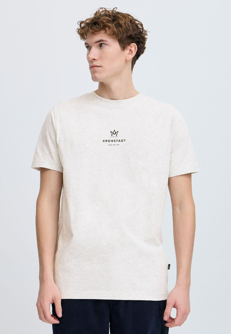 Camiseta beige clara hecha de tela suave, con un escote redondo y mangas cortas. En el frente presenta un logo negro con un diseño de corona.