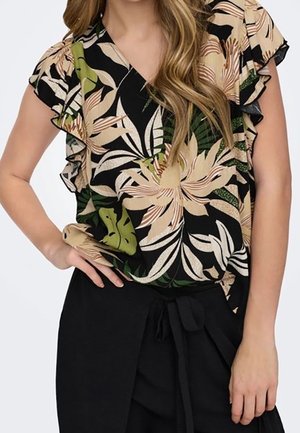Kvinde iført en kortærmet blomsterbluse med beige, grønne og sorte løvmønstre og sorte bukser med bindebælte i taljen.