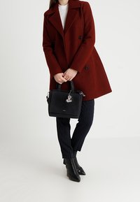 Svart läderhandväska med dubbla handtag, prydd med en silversmycke. Modellen bär en röd kappa, vit topp och rutiga byxor, tillsammans med svarta stövlar.