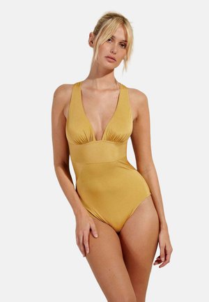 Donna bionda che indossa un costume intero giallo senape con scollo a V profondo su uno sfondo chiaro e semplice.