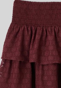 Jupe bordeaux avec une taille smockée, des volants superposés et un motif floral texturé. Le tissu présente un léger éclat.