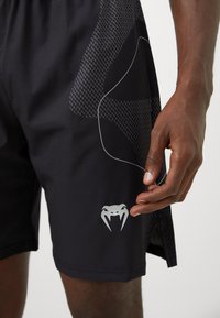 Nærbilde av en person som har på seg svarte treningsshorts med en sølvfarget logo med tenner nær kanten og subtile mønstre på stoffet.