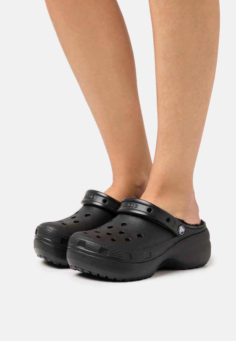 Sabots en plastique noir avec des trous de ventilation, un talon surélevé et une sangle sur le dessus. Surface lisse et fond texturé pour une meilleure adhérence.