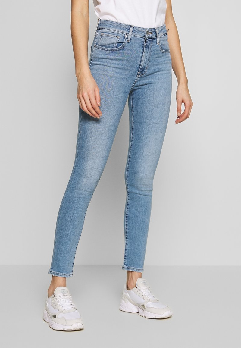 Jean skinny bleu clair avec une taille mi-haute, un léger délavage, cinq poches et des surpiqûres contrastantes, associé à des baskets blanches.