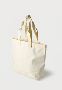 Τσάντα tote της Lacoste σε εκρού χρώμα με διπλές κοντές και μακριές λαβές, απλό υφασμάτινο σώμα και διακριτικό ανάγλυφο όνομα της μάρκας στα λουράκια.