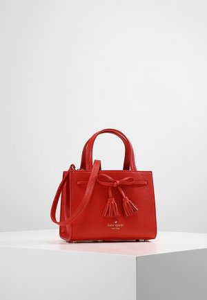 Czerwona skórzana torebka Kate Spade z górnymi uchwytami, odpinanym paskiem na ramię, ozdobą w formie kokardki i frędzlami, wystawiona na białym postumencie.