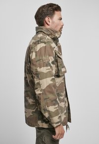 Kárová bunda vyrobená z odolnej tkaniny, s uzatváraním na zips, bočnými vreckami a nášivkami. PrevDominujúce zelené a hnedé odtiene.