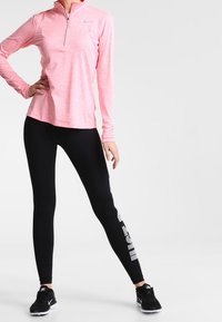 Jasnoróżowa bluza sportowa z długim rękawem i suwakiem na ćwierć, w połączeniu z czarnymi legginsami z białym logo Nike po boku. Czarne trampki.