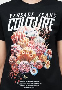 Μαύρο μπλουζάκι με το κείμενο «Versace Jeans Couture», ζωντανή γραφική απεικόνιση κοραλλιογενούς υφάλου και κοχυλιών, και κείμενο με θέμα την αγάπη κάτω από το νερό στο κάτω μέρος.