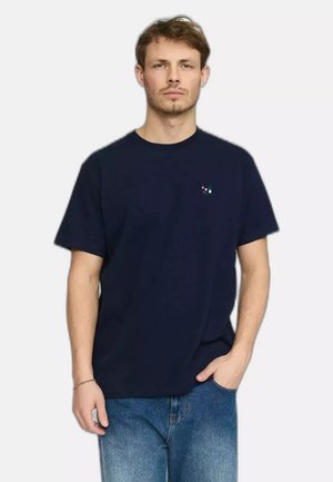 Jeune homme aux cheveux courts et bruns portant un t-shirt bleu marine avec un petit logo et un jean bleu, debout devant un fond uni.