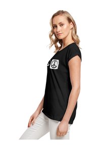 Schwarzes Baumwoll-T-Shirt mit zwei weißen Friedenssymbolen auf der Vorderseite, rundem Halsausschnitt und kurzen Ärmeln. Getragen mit weißen Hosen.
