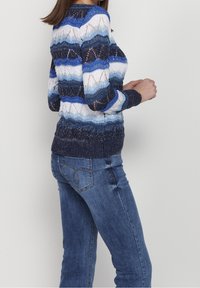 Femme portant un pull en tricot à rayures bleues, blanches et noires et un jean bleu, debout de profil avec les mains près de la poitrine devant un fond uni.