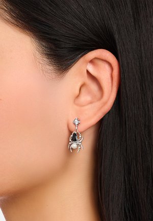 Ohr mit silbernem, spinnenförmigem Ohrring mit schwarzen und weißen Steinen und einer Perle, vor dunklem, glattem Haar.