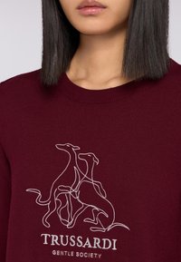 Weinroter Sweatshirt mit strukturiertem Strick, Rundhalsausschnitt und einer weißen Umrissillustration von zwei Hunden über dem Text "Trussardi Gentle Society".