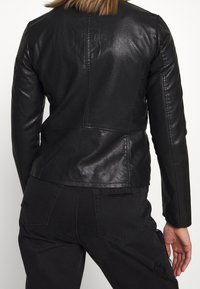 Veste en cuir noire au design ajusté, avec des coutures visibles et une texture lisse. Détails comprenant une taille légèrement cintrée et des manches longues.