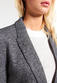 Veste grise texturée avec un revers net, portée sur un pull blanc présentant un motif en maille avec de petits points marron.