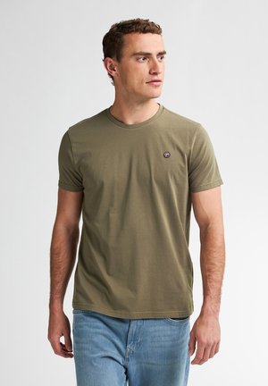 Man in olijfgroene korte mouwen t-shirt met klein rond logo op de borst, draagt lichtblauwe spijkerbroek, kijkt naar rechts tegen een effen lichte achtergrond.