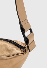 Metallic guld taske med en blød tekstur, som har en sort lynlås og en justerbar rem med plastikhardware detaljer.