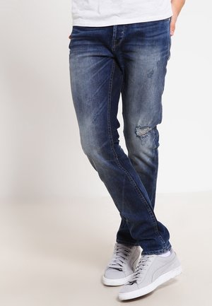 Person iført slanktsiddende blå slidte jeans og lysegrå sneakers, stående med krydsede ben mod en ensfarvet baggrund.