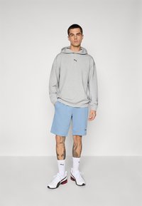 Mand iført en lys grå hættetrøje, blå shorts, hvide Puma-sokker og hvide sneakers, stående mod en ensfarvet lys baggrund.
