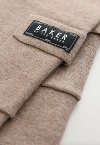 Beżowy sweter z dzianiny o teksturowanym wykończeniu, z czarną metką, na której w białych literach napisano "BAKER by TED BAKER", przyszytą na rękawie.