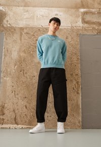 Vaaleansininen collegepaita, jossa pieni logo, mustat löysät housut ja valkoiset korkeavartiset tennarit. Kangas on sileää ja yksinkertaisen mallinen.