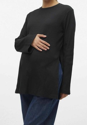 Persoon draagt een zwart shirt met lange mouwen en een blauwe spijkerbroek, hand rustend op de buik, torso zichtbaar, neutrale achtergrond.