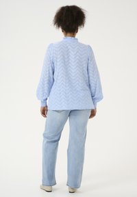 Chemisier bleu clair, transparent, avec un motif en zigzag, encolure froncée et manches bouffantes, associé à un jean clair et des chaussures beiges. Vue de dos.
