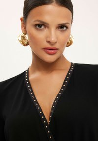 Czarna odzież z głębokim dekoltem w serek, ozdobiona złotymi ćwiekami wzdłuż dekoltu oraz dużymi złotymi kolczykami w kształcie kół. Gładka faktura materiału.
