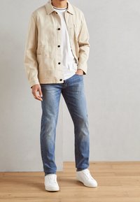 Chaqueta vaquera beige claro con botones negros, llevada sobre una camisa blanca. Jeans azul claro y zapatillas blancas completan el conjunto.
