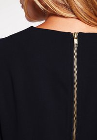 Černé šaty s detailem zlatého zipu na zadní straně. Tkanina vypadá hladce a má strukturovaný vzhled, což zvyšuje celkový čistý design.