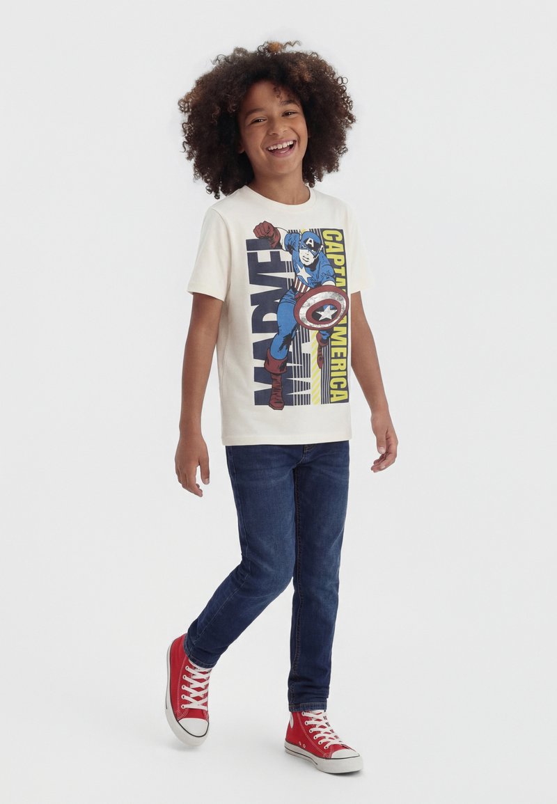 Uśmiechnięte dziecko spacerujące, ubrane w kremową koszulkę Captain America, ciemnoniebieskie dżinsy i czerwone trampki za kostkę na jednolitym białym tle.