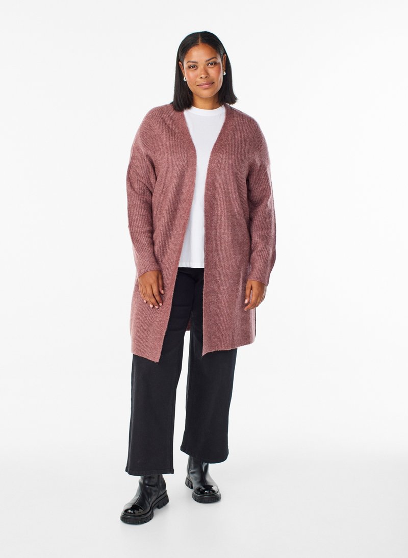 Donna dalla pelle media e capelli lisci indossa un lungo cardigan color malva, camicia bianca, pantaloni neri e stivaletti neri, in piedi su sfondo bianco.